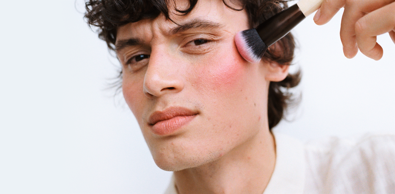 Nærbillede af en person, der påfører pink blush på kinden med en makeupbørste.