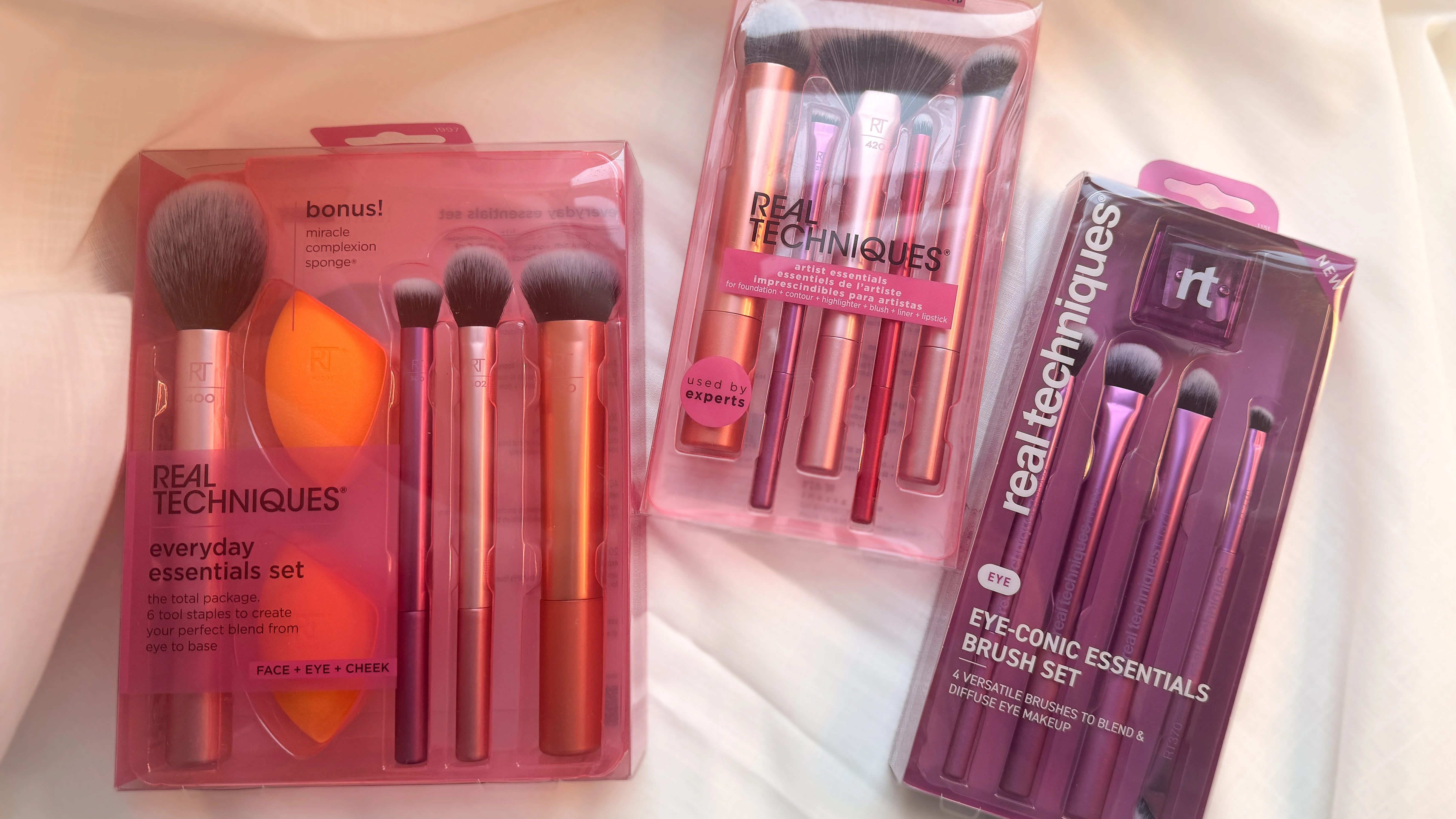 Tre Real Techniques makeupbørstesæt: Everyday Essentials med makeupsvamp, Artist Essentials til fuld makeup, og Eye-Conic Essentials med øjenbørster.