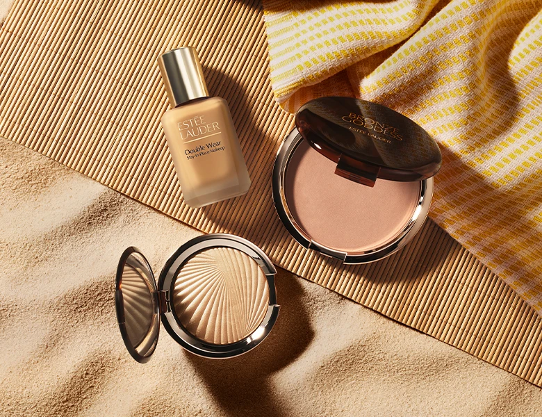 Her ses et billede med makeup-produkter fra Estée Lauder placeret i sand. Der ses blandt andet foundationen “Double Wear” samt bronzer- og pudderprodukter i kompakter.