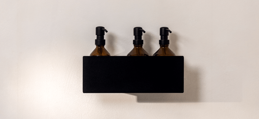 En enkel og stilren badevægsopstilling. På en sort, vægmonteret hylde står tre ens brune pumpeflasker i glas med sorte pumper, placeret side om side. Flaskerne er delvist skjult af hyldens front, så kun den øverste del med pumperne er tydeligt synlig. Baggrunden er en glat, lys beige væg, som skaber en rolig kontrast til den mørke hylde. Udtrykket er minimalistisk og organiseret.