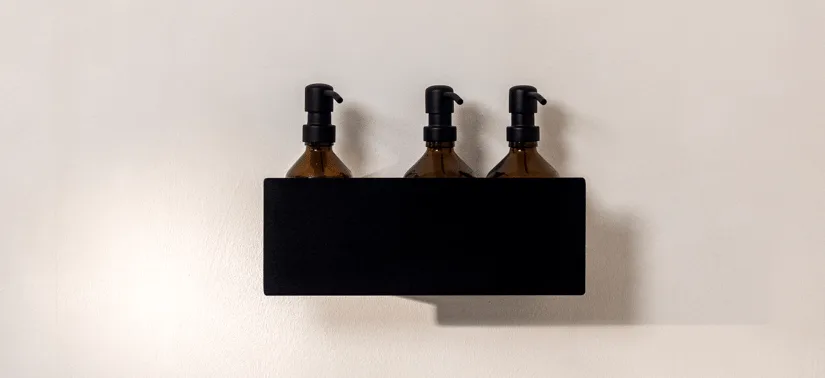En enkel og stilren badevægsopstilling. På en sort, vægmonteret hylde står tre ens brune pumpeflasker i glas med sorte pumper, placeret side om side. Flaskerne er delvist skjult af hyldens front, så kun den øverste del med pumperne er tydeligt synlig. Baggrunden er en glat, lys beige væg, som skaber en rolig kontrast til den mørke hylde. Udtrykket er minimalistisk og organiseret.