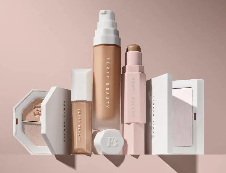 Her ses en beigebaggrund med forskellige Fenty Beauty produkter til ansigtet.