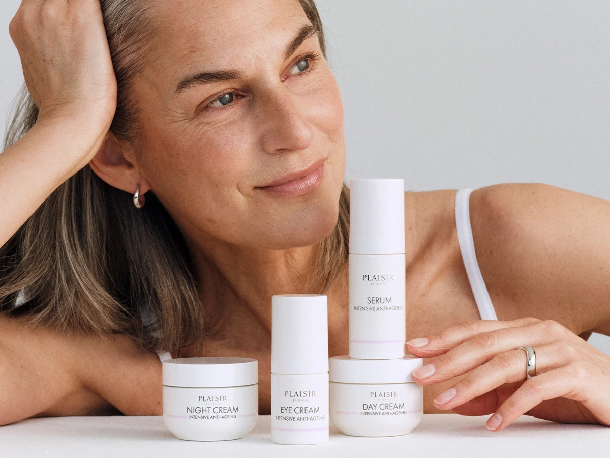 En kvinde viser fire hvide PLAISIR anti-ageing hudplejeprodukter: Natcreme, øjencreme, serum og dagcreme. Hun smiler let.