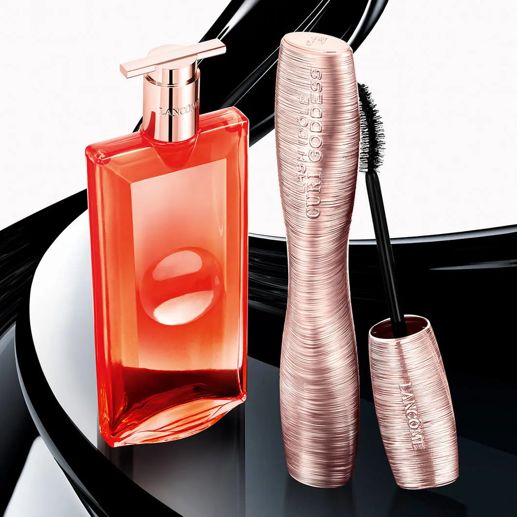Et produktbillede viser en rosa metallic Lancôme Lash Idôle-mascara ved siden af en orange Lancôme-parfumeflaske med skinnende låg. Mascaraen er åben, så den buede mascarabørste er synlig. Produkterne står foran sorte, bølgende mascarastrøg, som skaber et dramatisk og elegant beautyudtryk med fokus på makeup og luksusduft.