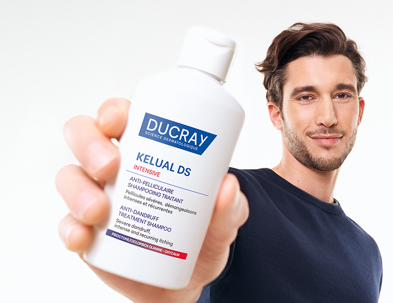 Her ses en mand, der holder en Ducray Kelual DS shampoo foran en lys baggrund.