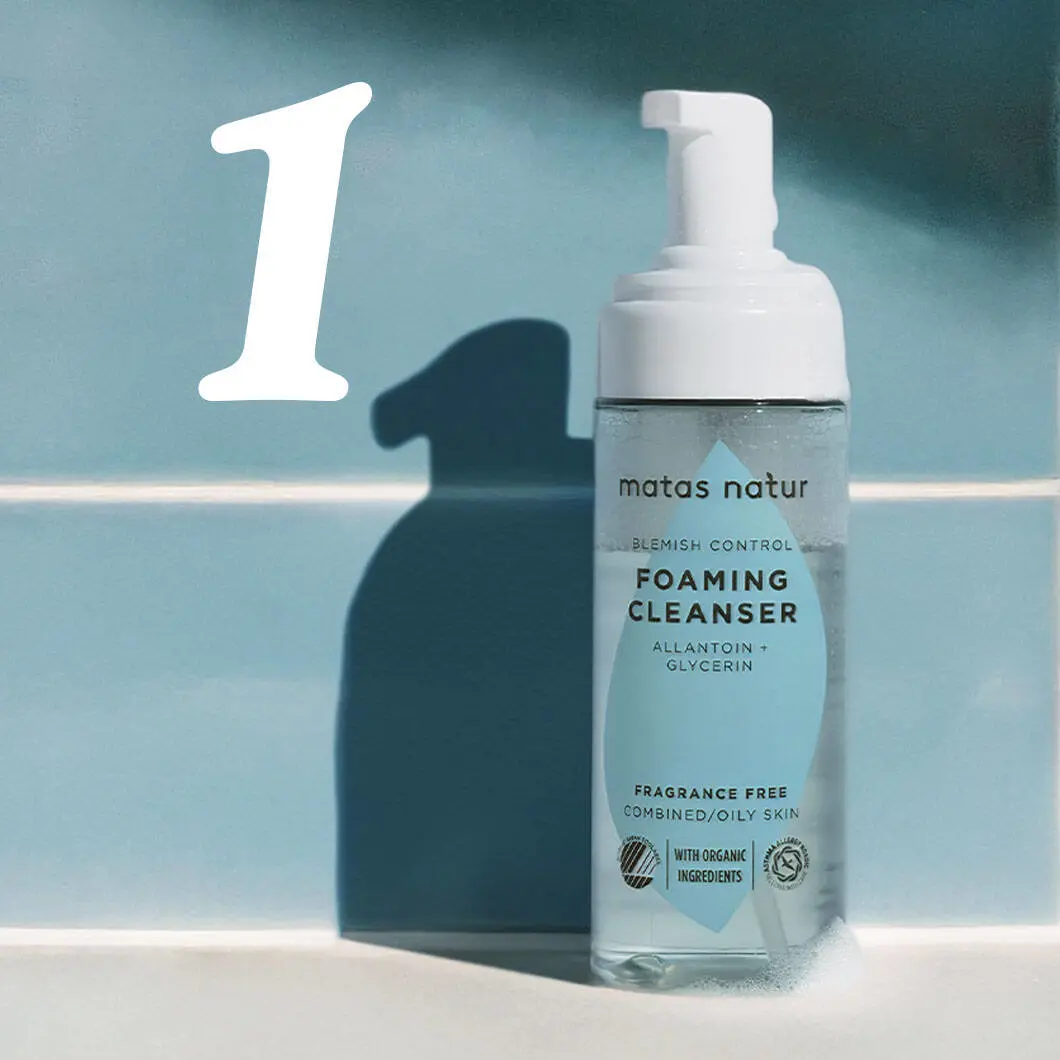 En flaske Matas Natur Blemish Control Foaming Cleanser står på en lys overflade mod en blå baggrund. Flasken er gennemsigtig med pumpe og indeholder en klar gel. På etiketten står blandt andet ‘Foaming Cleanser’, ‘Allantoin + Glycerin’ og ‘Fragrance Free’. Til venstre ses et stort hvidt tal 1, som indikerer første trin i en hudplejerutine. Flasken kaster en tydelig skygge bag sig.