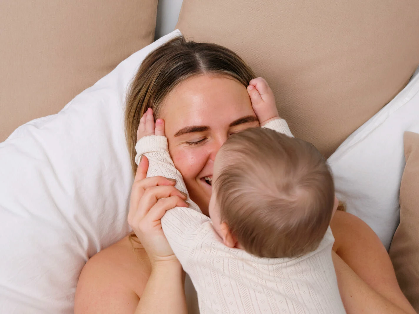 Mor og baby ligger tæt i en seng med hvide dyner og beige puder, mens babyen holder om morens ansigt og de smiler til hinanden.