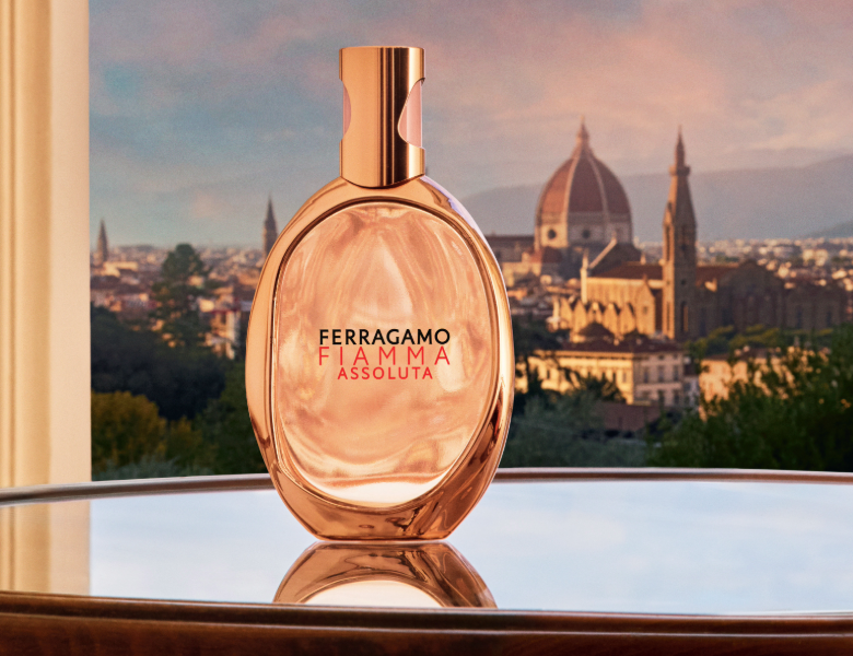 Her ses et billede af en parfumeflakon fra Ferragamo placeret foran en byudsigt ved solnedgang. På flakonen står teksten “FERRAGAMO FIAMMA ASSOLUTA”. Flakonen er i varme rosagyldne nuancer med guldfarvet låg.