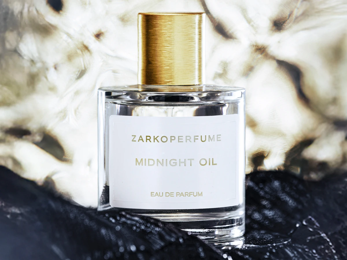 Her ses Zarkoperfume Midnight Oil Eau de Parfum i en transparent parfumeflakon med guldfarvet låg og hvid etiket. Flakonen står foran lyst og mørkt tekstil.