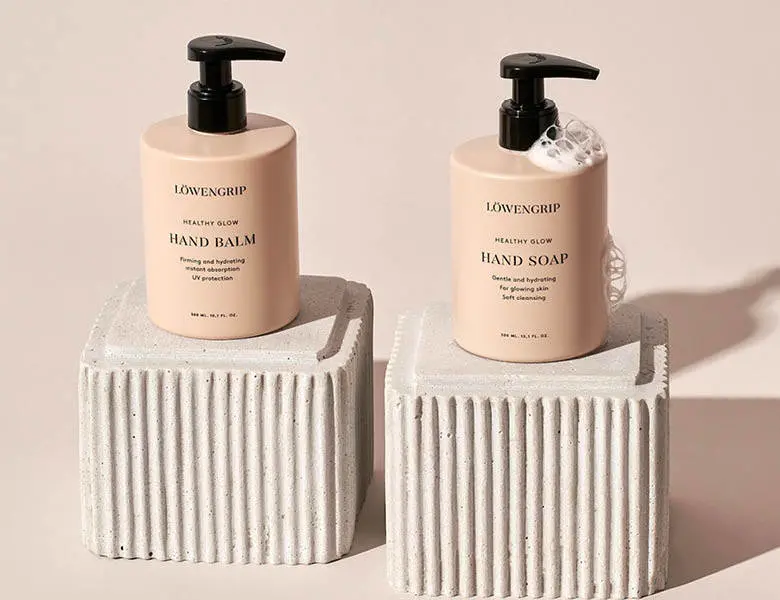 Her ses et billede med to produkter fra Løwengrip placeret på hver sin riflet blok mod en lys baggrund. På produkterne står teksten “HAND BALM” og “HAND SOAP”. Produkterne er i beige emballage med sorte pumper.