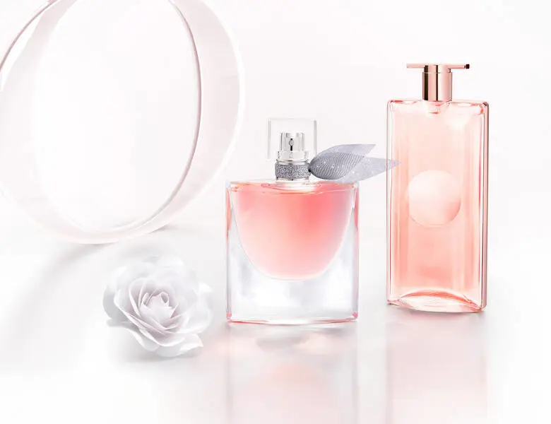 Her ses et billede med to parfumeflakoner i lyserøde nuancer placeret på en lys, reflekterende overflade mod en lys baggrund. Den ene flakon har en sløjfedetalje ved halsen. Ved siden af flakonerne ses en hvid blomst samt dekorative, runde elementer i baggrunden.