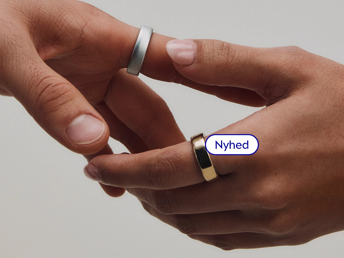 To hænder, den ene med en sølvfarvet Oura ring, den anden med en guldfarvet Oura ring. Tekst: Nyhed.