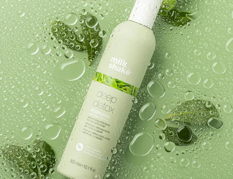 Her ses et billede af en shampoo-flaske fra milk_shake placeret mod en grøn baggrund med vanddråber og blade. På flasken står teksten “milk_shake deep detox shampoo”. Flasken er i lysegrøn og hvid emballage.