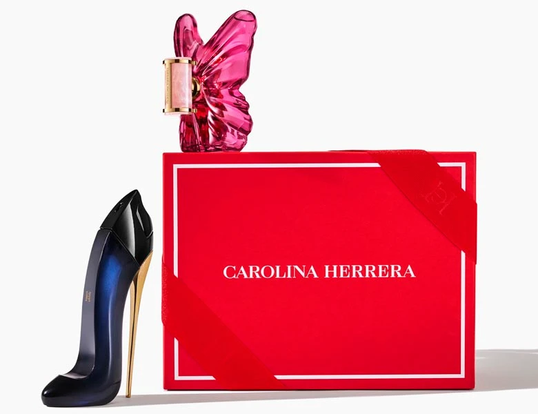 Her ses et billede med to parfumeflakoner placeret ved siden af en rød æske med teksten “CAROLINA HERRERA”. Den ene flakon er formet som en højhælet sko i mørkeblå med guldfarvet hæl, og den anden har en pink, gennemsigtig form.
