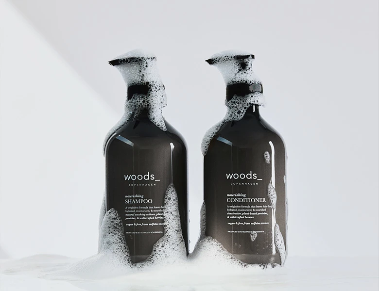 Her ses WOODS_ shampoo og conditioner i mørke flasker med skum på en lys baggrund.