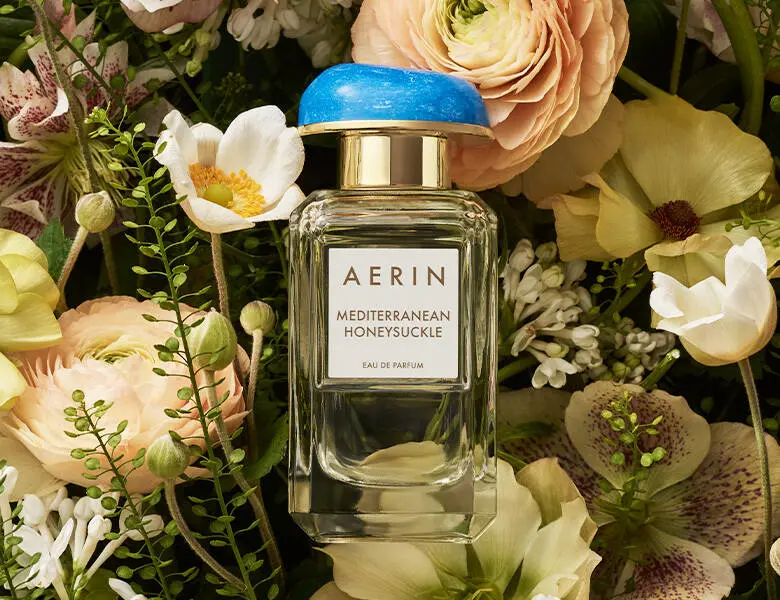 Her ses AERIN Mediterranean Honeysuckle Eau de Parfum i en transparent flakon med blåt låg og guldfarvede detaljer, omgivet af lyse blomster og grønne blade.