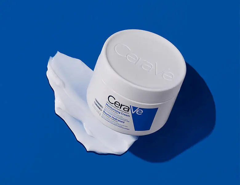 En rund, hvid krukke fra CeraVe står på en blå baggrund. Låget har præget logo, og krukken står oven på en udtværet mængde hvid creme, som breder sig ud under den. Billedet har fokus på produktets tekstur og en ren, klinisk æstetik.