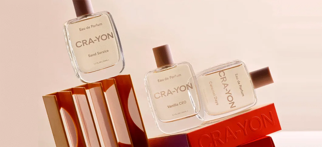 Her ses tre CRA-YON Eau de Parfum-flakoner med duftene Sand Service, Vanilla CEO og Caramel Days. Flakonerne er arrangeret på orange og røde dekorative elementer mod en lys rosa baggrund.