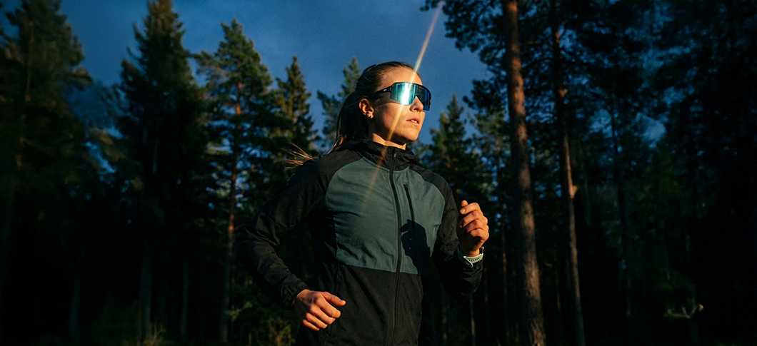 Kvinde løber i en skov ved lavt sollys og har store sportsbriller på samt mørkt løbetøj fra Ortho Movement