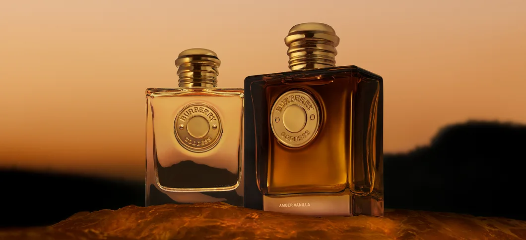 Her ses et billede med to parfumeflakoner fra Burberry Goddess placeret mod en baggrund med varme solnedgangsfarver. Den ene flakon er klar, mens den anden har en mørkere ravfarve med teksten “Amber Vanilla”.