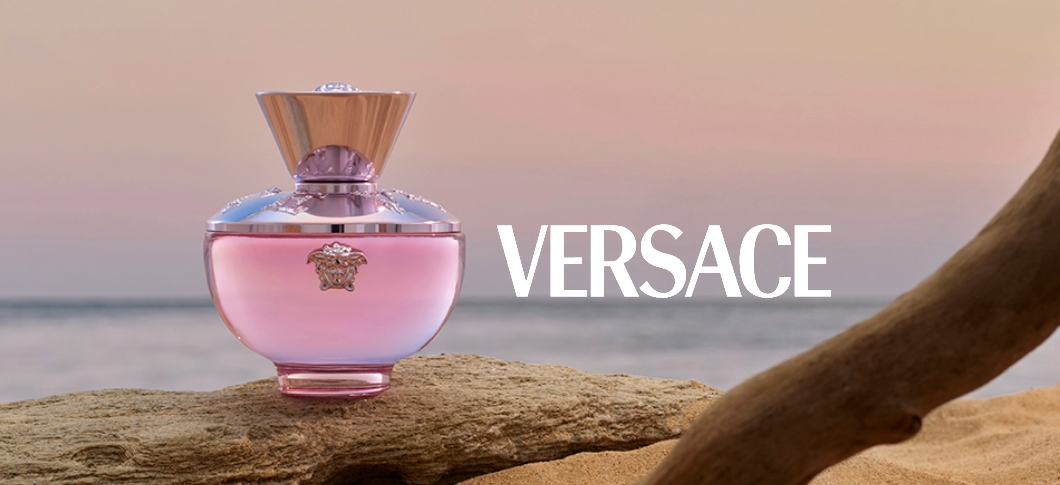Her ses en Versace Dylan parfume i en pink flakon stående på en sten og med havet som baggund med en lyserød himmel.