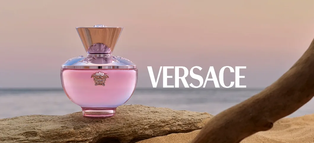 Her ses en Versace Dylan parfume i en pink flakon stående på en sten og med havet som baggund med en lyserød himmel.
