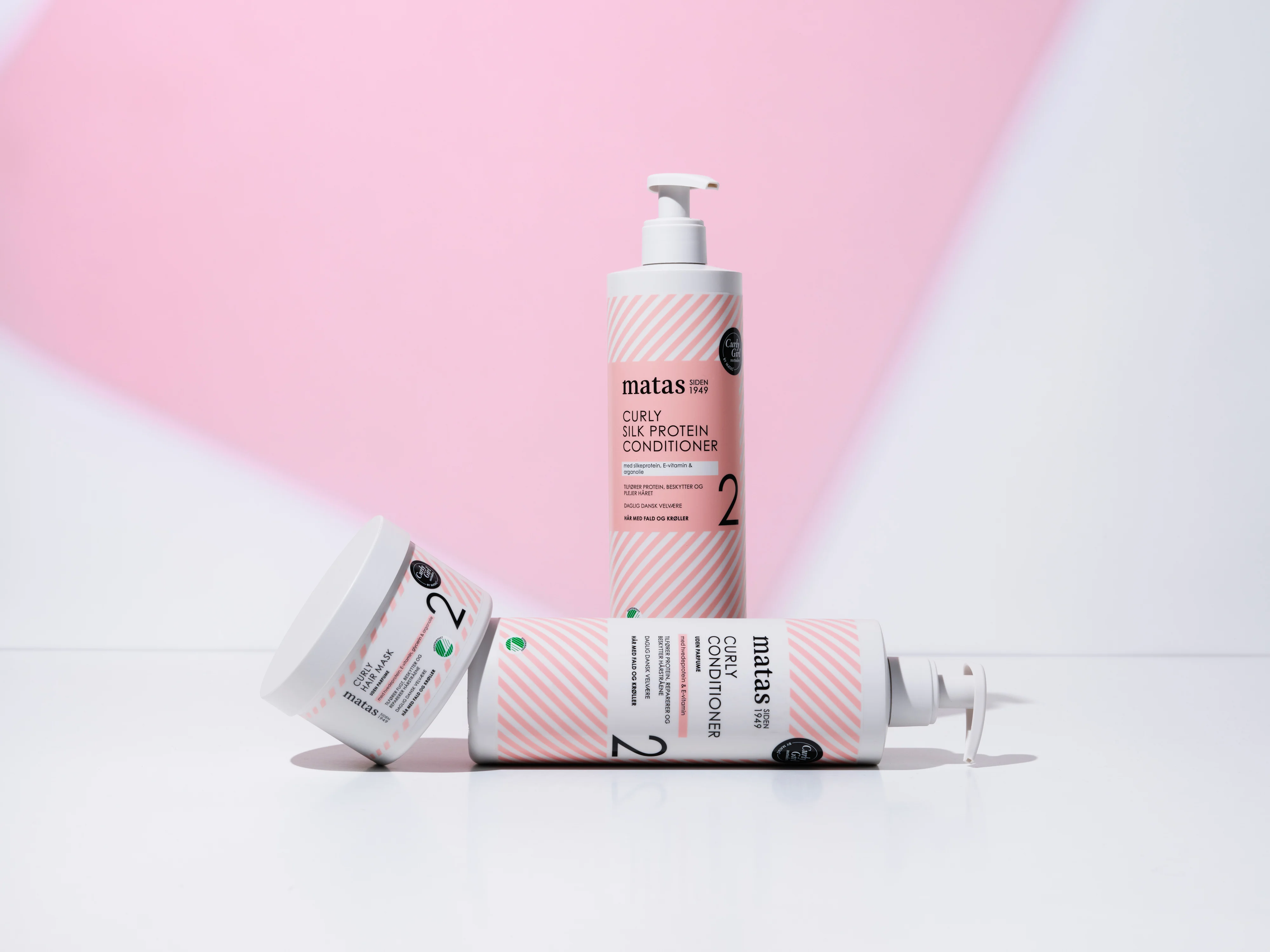 Her ses Matas Striber Curly Silk Protein Conditioner, Curly Conditioner og Curly Hair Mask på en lys baggrund med et lyserødt grafisk farvefelt.