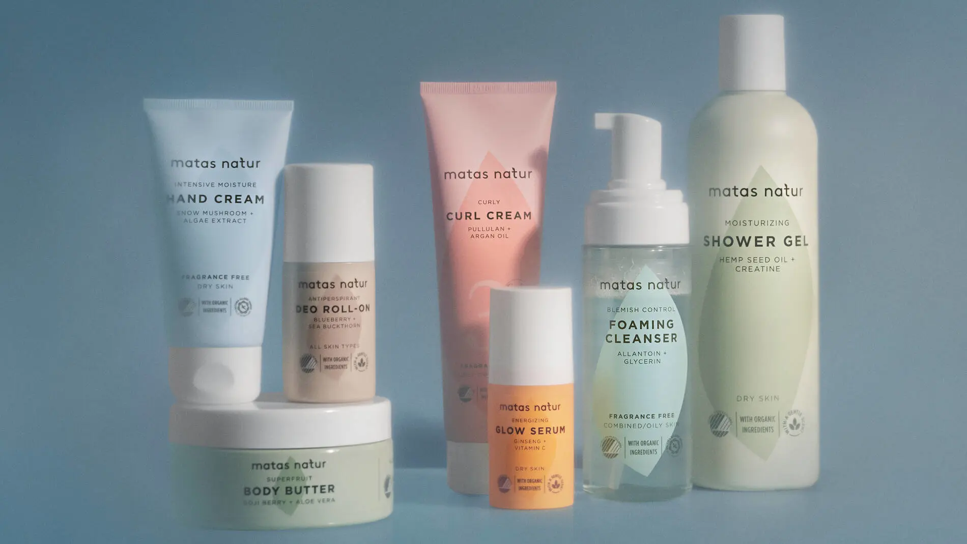 Et udvalg af produkter fra Matas Natur står opstillet foran en lyseblå baggrund i blødt lys. Produkterne består blandt andet af håndcreme, deodorant roll-on, curl cream, glow serum, foaming cleanser, shower gel og body butter i pastel-farvede emballager i blå, grønne, rosa og gule nuancer. Nogle produkter står direkte på overfladen, mens andre er placeret oven på en rund krukke. Emballagerne har enkle former og rolige farver, som giver billedet et rent og naturligt udtryk.