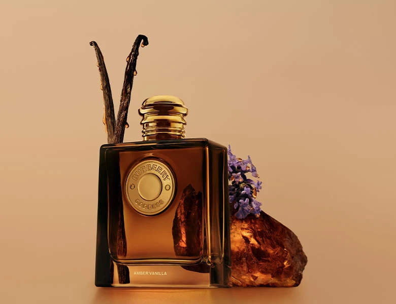 Her ses et billede med en parfumeflakon fra Burberry Goddess placeret foran en ravfarvet sten sammen med vaniljestænger og lilla blomster mod en varm beige baggrund. På flasken står blandt andet “Amber Vanilla”.
