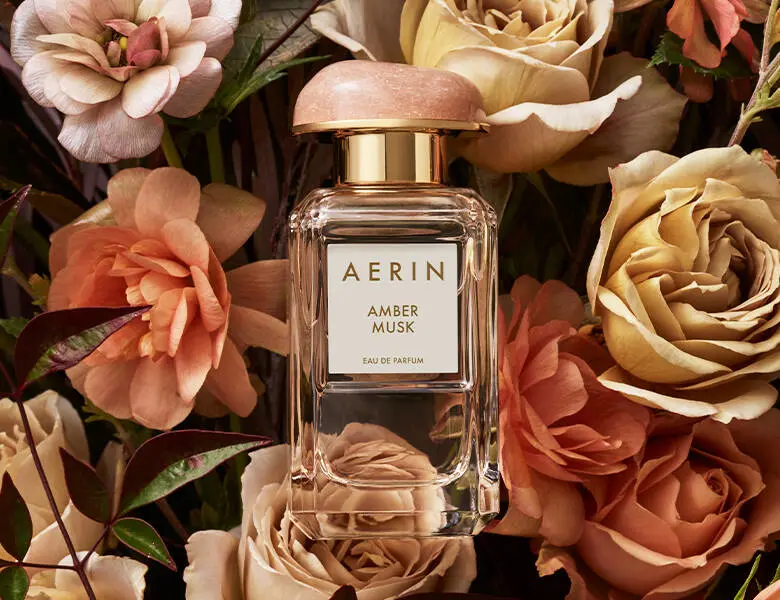 Her ses AERIN Amber Musk Eau de Parfum i en transparent flakon med rosa låg og guldfarvede detaljer, omgivet af blomster i rosa, fersken og cremefarvede nuancer.