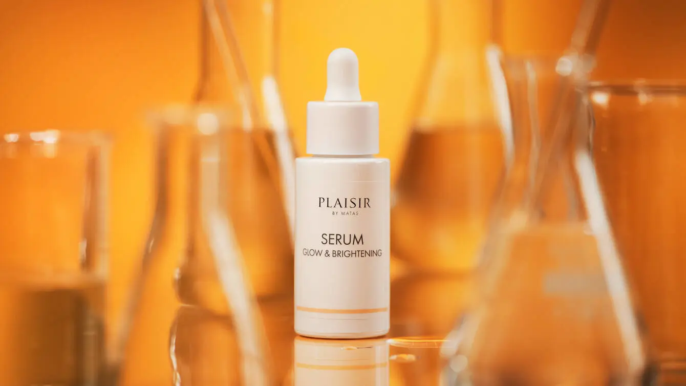 Produktbillede af “Plaisir by Matas Serum Glow & Brightening” på en varm orange baggrund. I midten står en hvid serumflaske med pipette, omgivet af flere glasbeholdere med væske, som er sløret i baggrunden. Flasken står på en blank overflade, der spejler produktet. Billedet har et varmt og laboratorieinspireret udtryk med fokus på aktiv hudpleje og glødgivende serum.