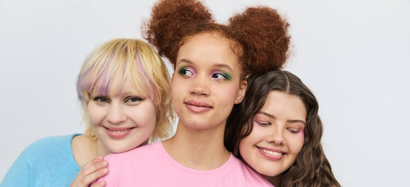 Her ses et billede med tre personer placeret tæt sammen mod en lys baggrund. De har forskellige frisurer og makeup i farverige nuancer og smiler mod kameraet.