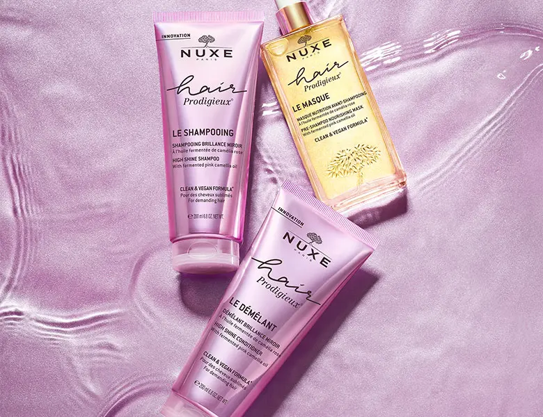 Tre Nuxe Hair Prodigieux produkter: En pink shampoo, en guld hårmaske og en pink balsam på en lilla baggrund.