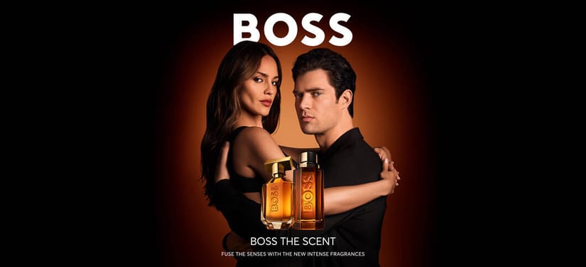Her ses et billede med to personer placeret centralt foran en mørk baggrund med orange lys. Øverst står teksten “BOSS”, og nederst står “BOSS THE SCENT” samt “FUSE THE SENSES WITH THE NEW INTENSE FRAGRANCES”. Foran personerne ses to parfumeflakoner i gyldne nuancer.