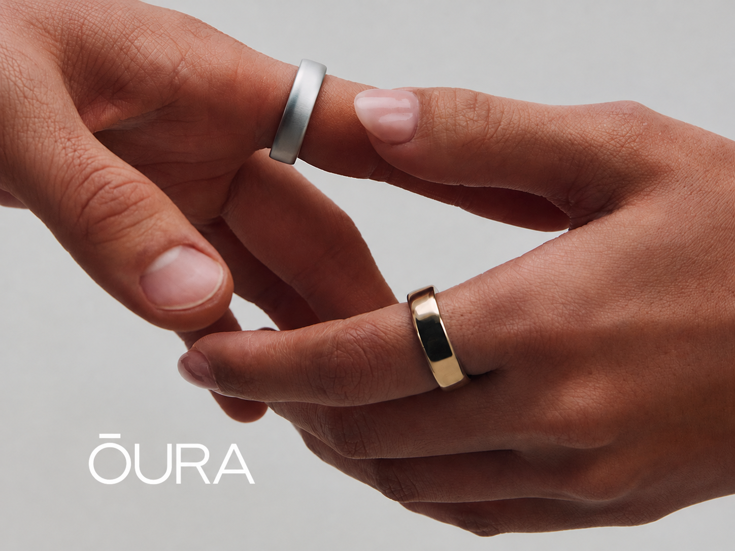 To hænder, den ene med en sølv Oura-ring og den anden med en guld Oura-ring, der rører hinandens pegefingre. Oura-logo.