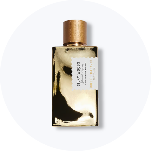 Her ses et billede af en parfumeflakon fra Goldfield & Banks placeret centralt mod en lys, rund baggrund. På flakonen står teksten “SILKY WOODS”. Flakonen har en guldfarvet, reflekterende overflade og et træfarvet låg.