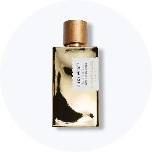 Her ses et billede af en parfumeflakon fra Goldfield & Banks placeret centralt mod en lys, rund baggrund. På flakonen står teksten “SILKY WOODS”. Flakonen har en guldfarvet, reflekterende overflade og et træfarvet låg.