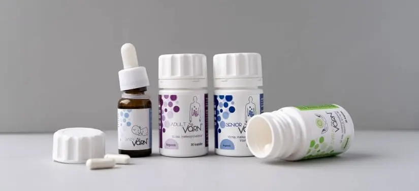 Banner med Värn kosttilskud: brun pipetteflaske til baby samt flere hvide bøtter med kosttilskud til børn, voksne og seniorer. Produkterne står på en lys overflade foran en neutral grå baggrund, og en åben bøtte med kapsler ligger foran.