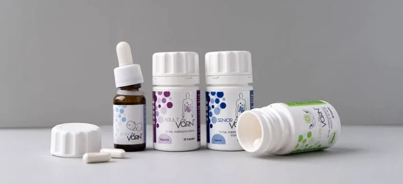 Banner med Värn kosttilskud: brun pipetteflaske til baby samt flere hvide bøtter med kosttilskud til børn, voksne og seniorer. Produkterne står på en lys overflade foran en neutral grå baggrund, og en åben bøtte med kapsler ligger foran.