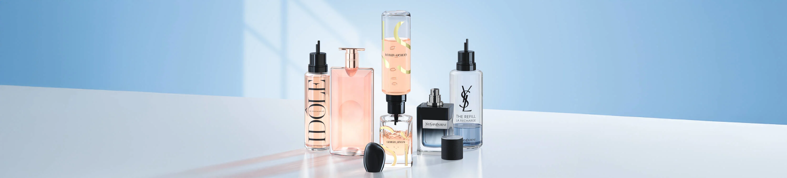 En række parfume- og beautyprodukter står samlet på en lys overflade foran en blå baggrund. Produkterne består af elegante glasflasker i forskellige former og størrelser med pumpe- og refillfunktioner. Flere af flaskerne er i lyserøde og gennemsigtige nuancer, og nogle har synlige slanger eller indsatser, der viser refill-systemet. Kendte mærker som Dior, Giorgio Armani og Yves Saint Laurent kan ses på emballagen. Billedet har et rent og moderne udtryk med fokus på genopfyldelige skønhedsprodukter.