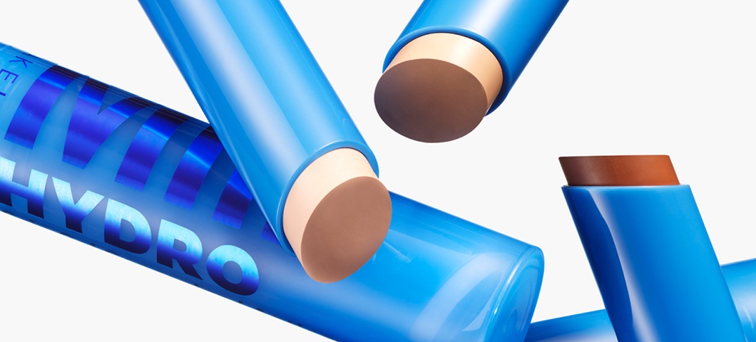 Her ses et billede med flere makeup-sticks i blå emballage placeret mod en lys baggrund. Produkterne vises både lukkede og åbne med synlige farver i beige og brun nuancer, og på emballagen står blandt andet “HYDRO”.