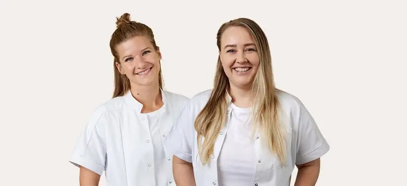 To smilende kvinder står side om side foran en lys baggrund. Begge bærer lyse uniformer, som minder om arbejdstøj inden for sundhed eller apotek. Den ene har håret sat op i en knold, mens den anden har langt blondt hår. Billedet har et venligt og professionelt udtryk.