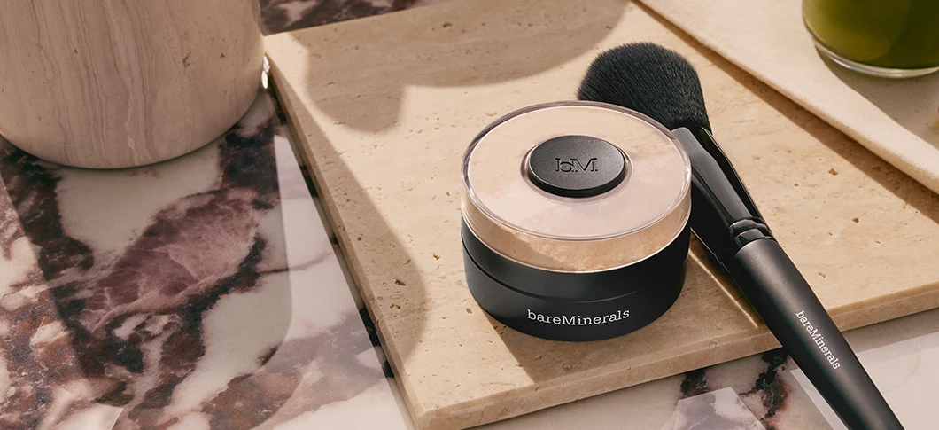 En bareMinerals løs pudderfoundation og en sort makeupbørste med teksten 'bareMinerals' på en lys stenplade og marmoroverflade.