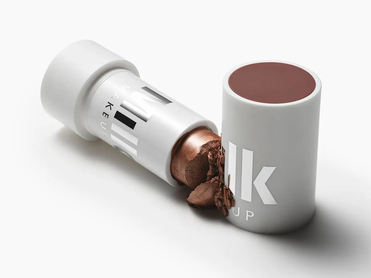 Her ses et åbent makeupstick fra Milk Makeup med en metallisk brun nuance. Produktet ligger ved siden af låget, og en smule af den cremede makeup er brudt af foran sticket.