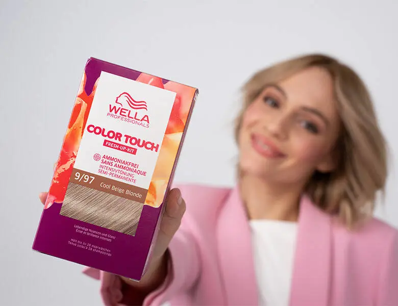 Her ses et billede af en person, der holder en hårfarveæske fra Wella Professionals frem mod kameraet. På æsken står teksten “COLOR TOUCH Fresh-Up-Kit” samt “9/97 Cool Beige Blonde”. Personen i baggrunden er iført pink blazer og smiler.