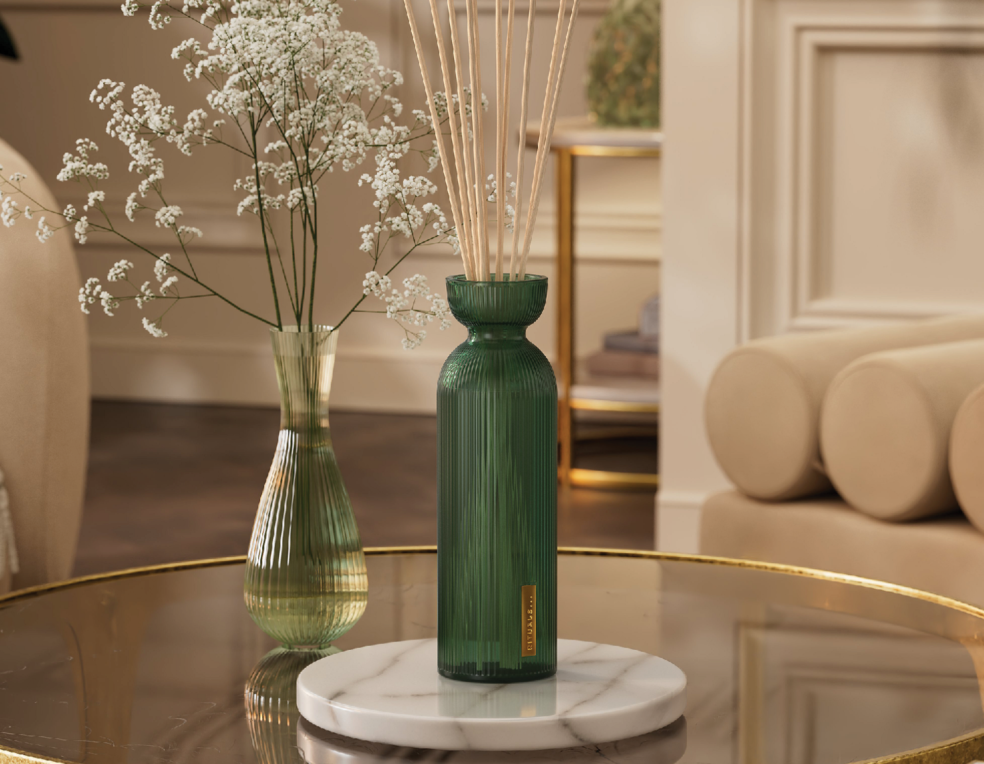 Grøn glasdiffuser med 'RITUALS...' etiket og pinde, ved en vase med hvide blomster på et glasbord.