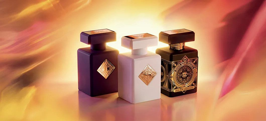 Her ses et billede med tre parfumeflakoner fra Initio Parfums Privés placeret mod en varm, gylden baggrund. Flakonerne er i mørkelilla, hvid og sort med guldfarvede detaljer, og den sorte flakon har teksten “OUD FOR GREATNESS” på fronten.