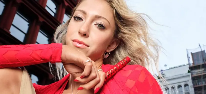 Her ses en kvinde med lyst hår og rød bluse, der holder en rød mascara. I baggrunden er der bygninger og blå himmel.
