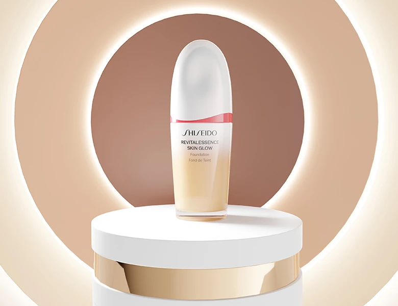Her ses et billede af et foundation-produkt fra Shiseido placeret på et podium mod en beige baggrund med cirkulært lys. På produktet står teksten “SHISEIDO REVITALESSENCE SKIN GLOW Foundation”.