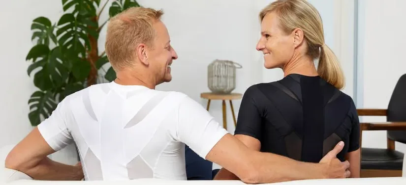 En mand i hvidt tøj og en kvinde i sort tøj begge med ryggen til som har Anodyne holdningskorrigerende tøj på. De kigger glade på hinanden